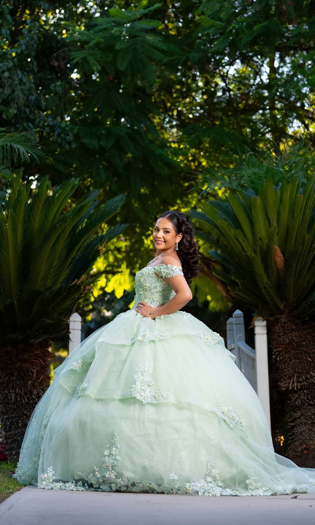 A young woman stands outdoors on a path surrounded by lush green foliage and palm-like plants, posing with one hand on her hip while wearing the Quinceanera Dress CHF3104 By Chicasโa sage green gown featuring a floral embroidered corset bodice.
